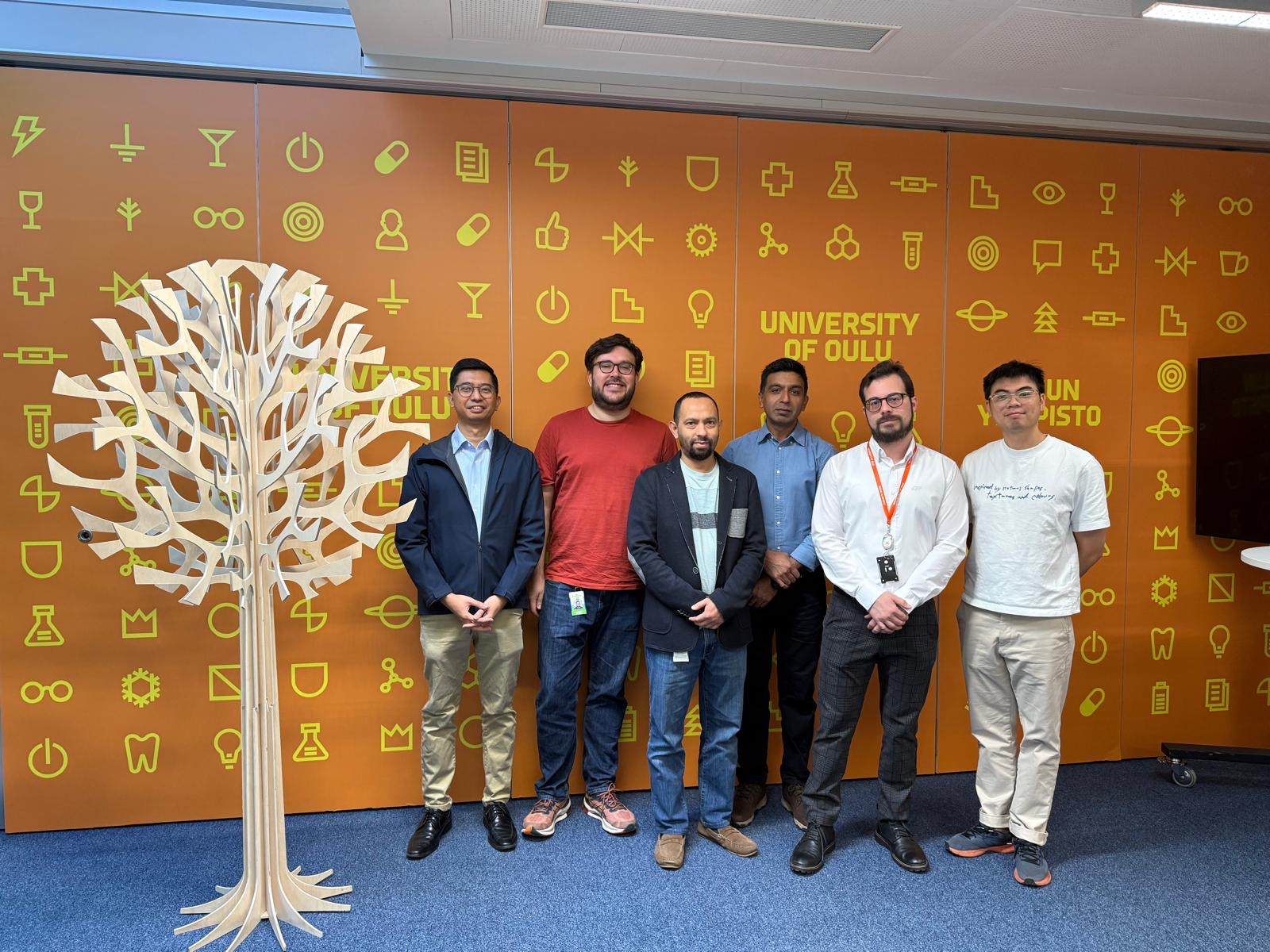 Group photo of six researchers in front of an orange University of Oulu wall, next to a wooden tree sculpture