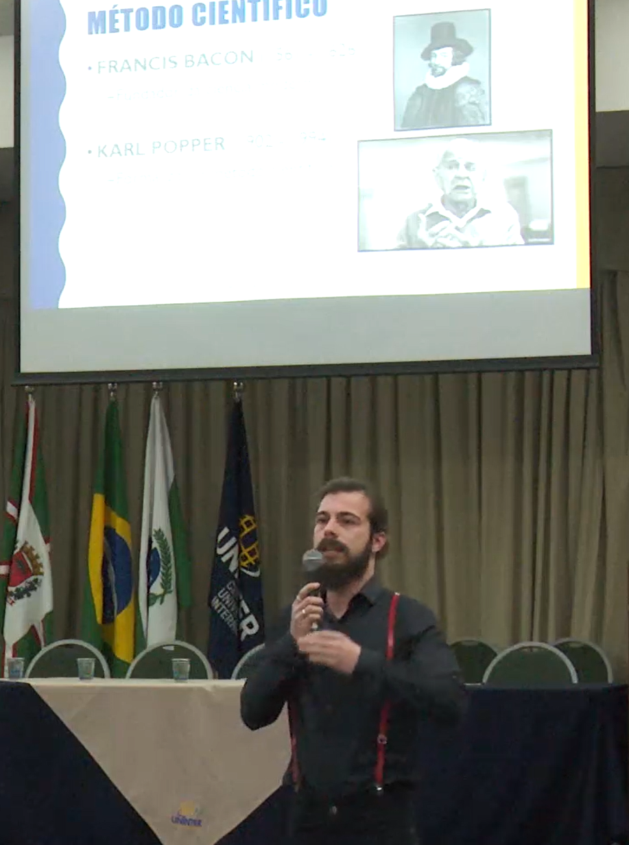 Speaking on stage in front of a slide about the scientific method, Francis Bacon and Karl Popper, with flags behind