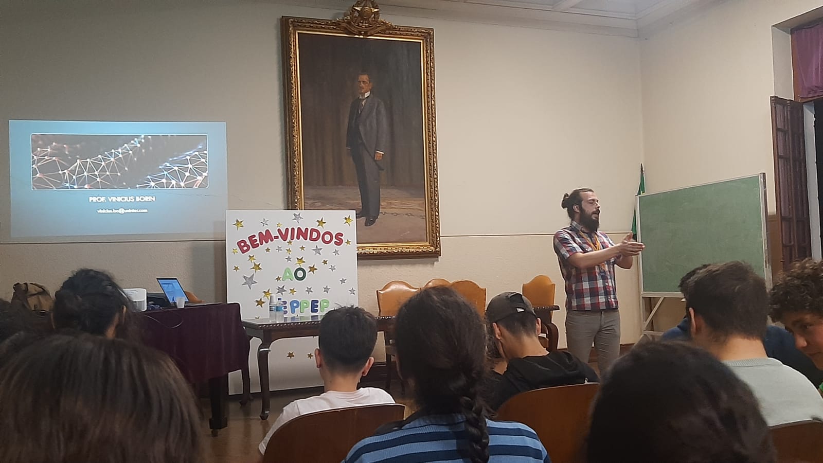 Speaking to a room of seated students beneath a portrait, next to a hand-decorated welcome sign